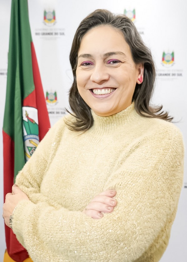 A imagem mostra uma mulher sorridente, de pele clara e cabelos castanhos escuros, lisos e na altura dos ombros. Ela está usando uma blusa de lã bege com brilho e maquiagem com sombra rosa. Seus braços estão cruzados e ela está olhando diretamente para a câmera. Ao fundo, há uma bandeira do estado do Rio Grande do Sul, parcialmente visível à esquerda da imagem, e um painel branco com o brasão e o texto “Governo do Estado do Rio Grande do Sul” repetido. A mulher transmite uma imagem de confiança e simpatia, possivelmente em um ambiente institucional.